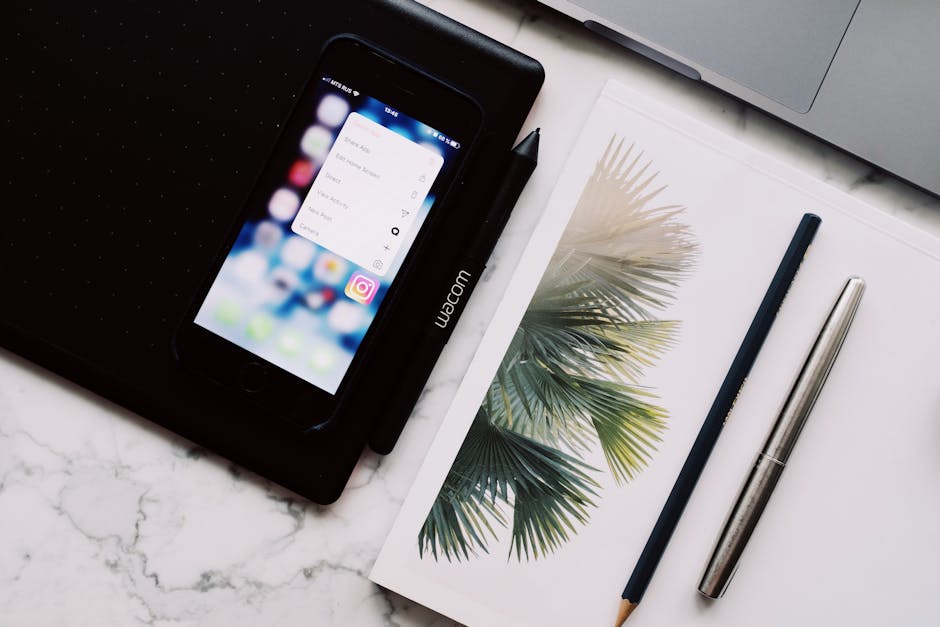 A close-up view of a modern workspace featuring a smartphone, Wacom drawing tablet, and writing tools on a marble surface.