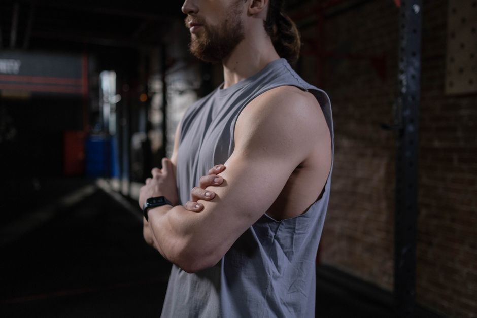 Confident man in a gray tank top standing cross-armed in a gym setting.