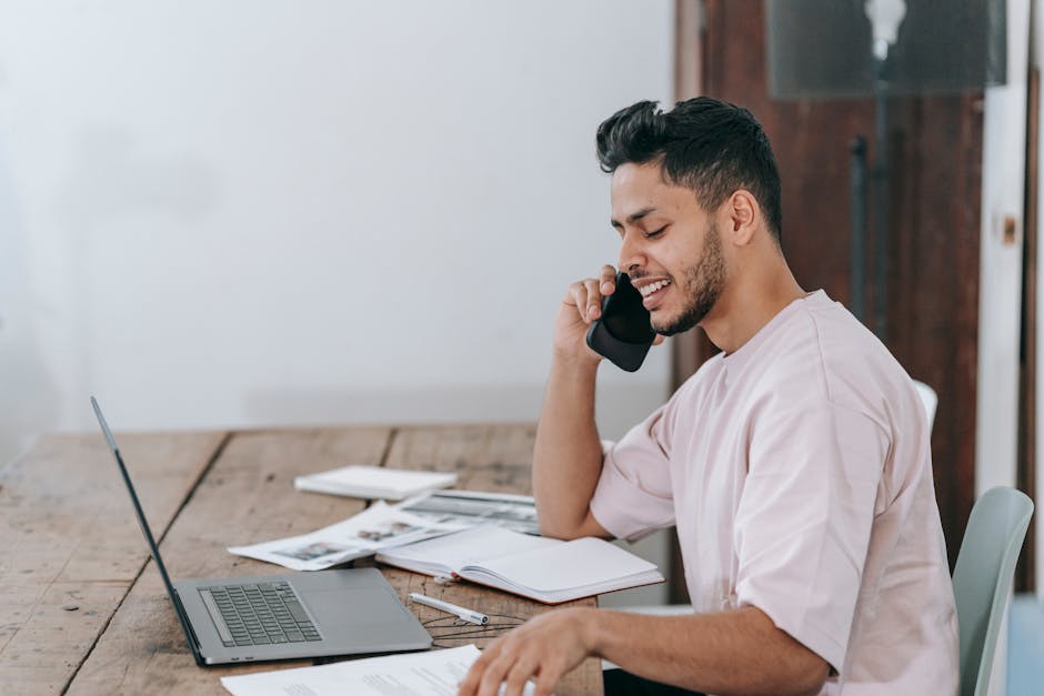 Side view of positive young Hispanic male entrepreneur in casual clothes checking documents and talking on smartphone while working remotely with laptop in home office